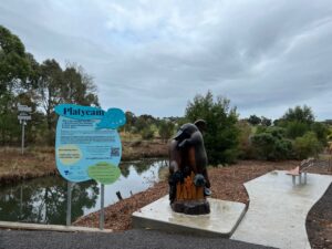 Image shows a river with a camera on a pole on thefar bank, and signage, a platypus sculpture and seating area.